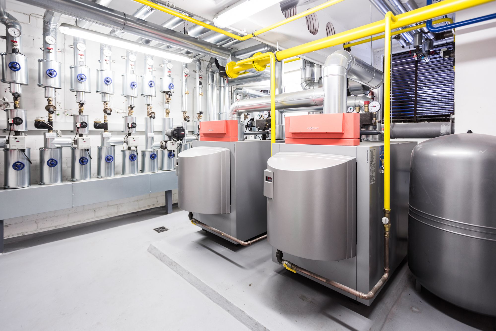 Industrieller Heizungsraum mit zwei großen grauen Heizkesseln und gelben Sicherheitsrohren